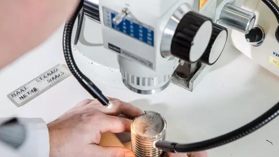 A lab technician positions a threaded metal sample under a stereomicroscope for macrographic examination