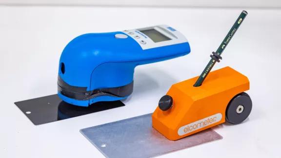 A handheld glossmeter and a separate pencil hardness tester positioned on metal test panels