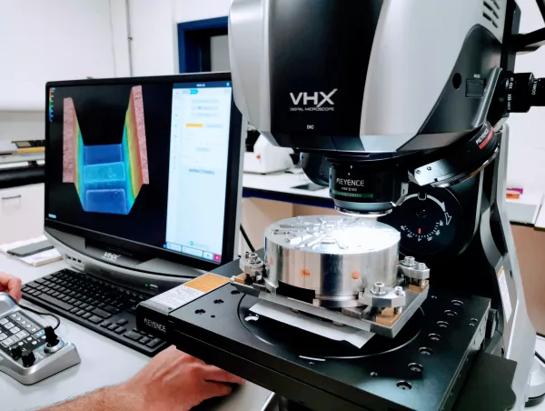 A Keyence VHX digital microscope measures a weld cross-section on a metal fixture, with the colour-coded profile shown on screen