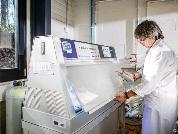 A lab technician inspects test panels with a handheld measuring device inside a QUV weathering chamber