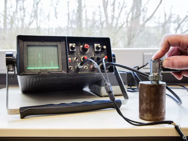 An analog ultrasonic flaw detector displays a reflected signal while a probe is held against a metal test block