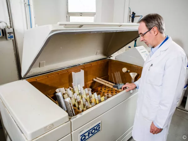 A lab technician inspects coated metal test panels loaded on the rack inside an open Q-Fog salt spray chamber