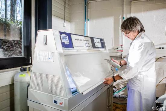 A lab technician inspects test panels with a handheld measuring device inside a QUV weathering chamber