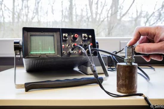 An analog ultrasonic flaw detector displays a reflected signal while a probe is held against a metal test block