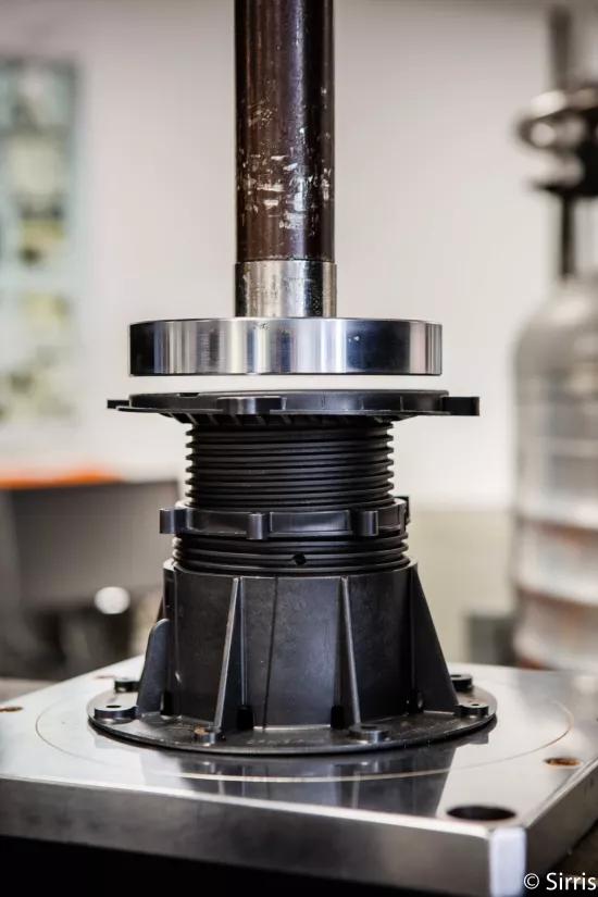 A test sample is positioned under the compression plate of a testing machine, ready for loading
