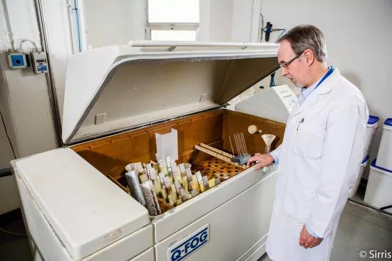 A lab technician inspects coated metal test panels loaded on the rack inside an open Q-Fog salt spray chamber