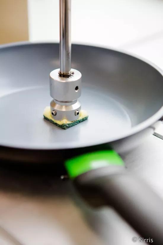 Close-up of a linear abraser test head fitted with a scouring pad, resting on a coated frying pan surface